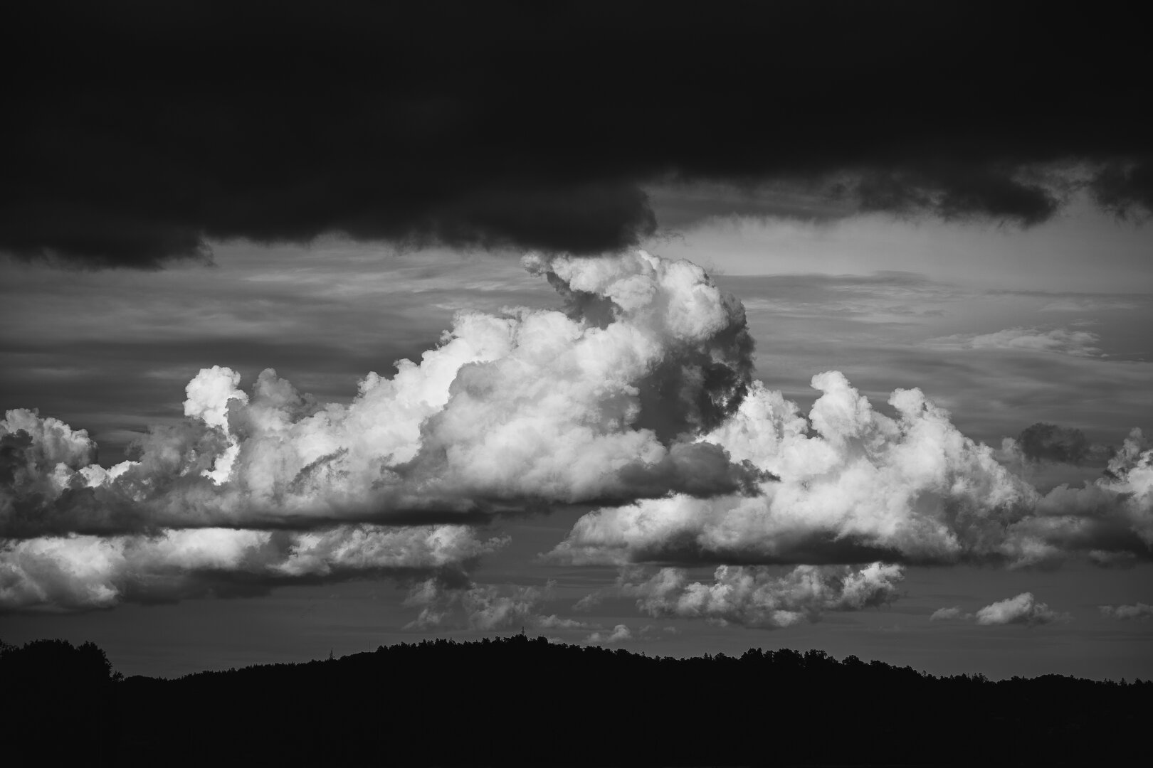 A monochrome greyscale high contrast photograph, of an undulating, hilly, forested landscape that is only appearing as a black silhouette along the bottom edge of the photo. The main feature of the photograph are the clouds.

Along the top edge is a band of very dark quite nearby clouds and between that band and the landscape silhouette is a dramatic, wide band of large cumulus clouds stretching toward the horizon.

The large fluffy clouds are white and sunlit on the top sides and dark on the bottom sides.

In the background sky are horizontal grey clouds that fill the sky much higher up.