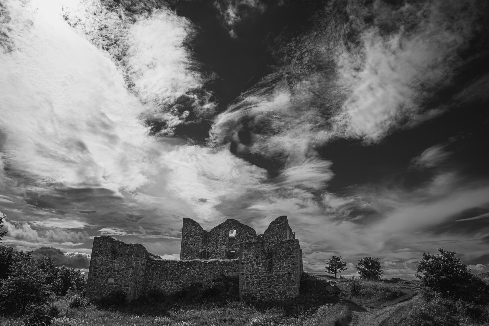 A wide angle monochrome photograph of the ruins of a large old stone house. The house sits in a hilly landscape with some trees, under half overcast sky that dominates the photo, with light clouds criss-crossing the sky and the sun lighting them from behind.

The house has the bases of what looks like two square tower in the foreground, with a wall running between them, with the house itself a bit further back. The house has two rows of large windos, with the wall broken and jagged along the second row of windows and the roof entirely missing.