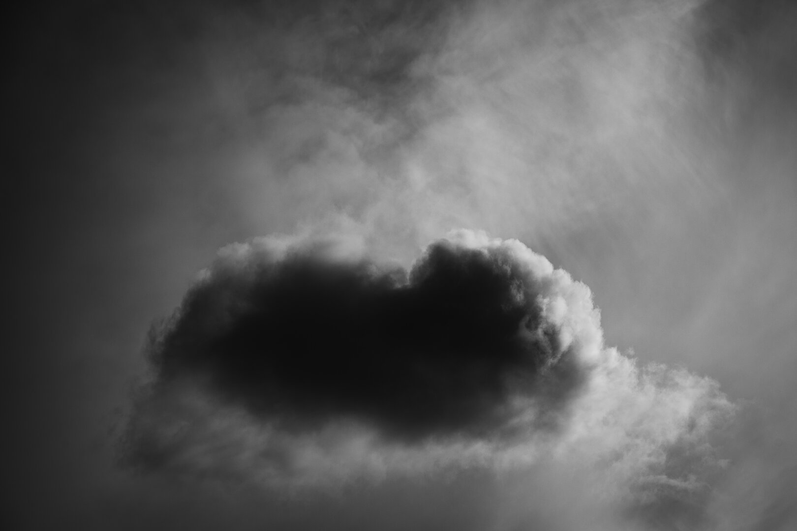 A high contrast monochrome photograph of a very small cloud. The rounded, slightly oblong and fluffy floud is in the lower half of the frame and is very dark in the center and light, almost white, along the fluffy edge, backlit by the sun.

In the background the sky is covered by more, and more distant clouds, thin and sparse enough to let some sunlight through.
