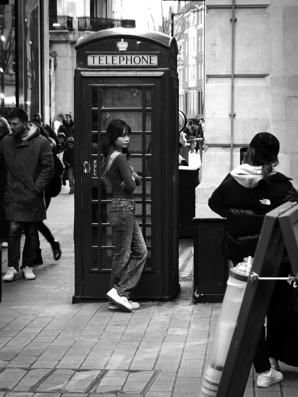Black and white. A dark-haired young woman wearing jeans and a long-sleeved t-shirt leans against the door of a London telephone box while her friend takes a photo with her phone. Black and white. A dark-haired young woman wearing jeans and a long-sleeved t-shirt leans against the door of a London telephone box while her friend takes a photo with her phone.
