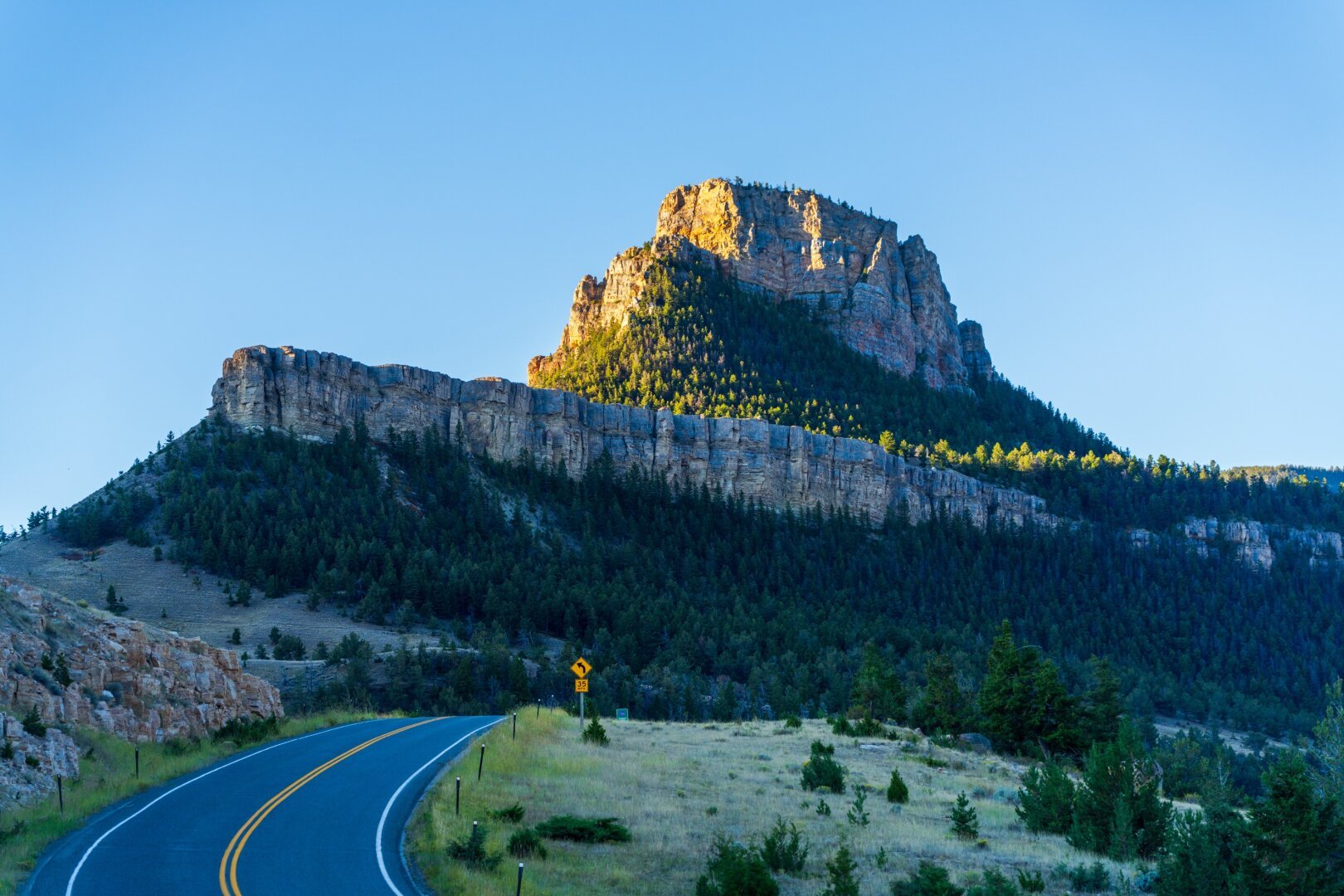 A large rock formation sits sun-lit on a big sharp bluff over a highway. [Sony A7R V / Tamron 25-200]