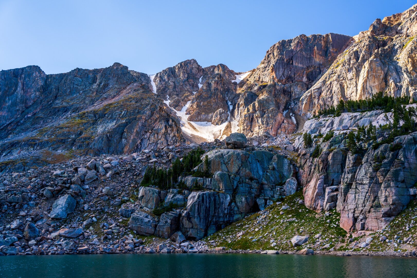 A rocky mountainside looms over a blue lake, with all sorts of colors on the rocks, and snow in bits. [Sony A7R V / Tamron 25-200]