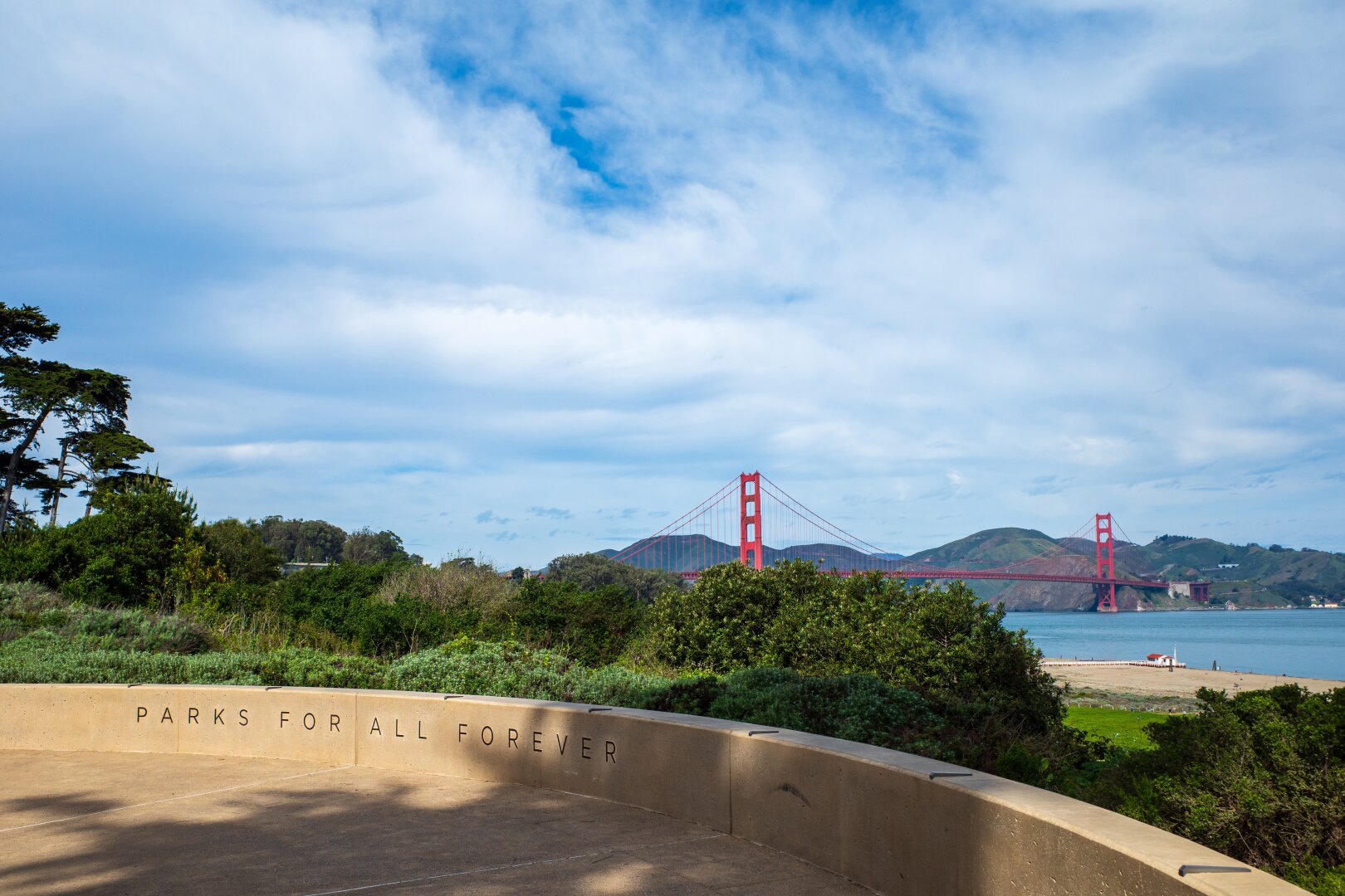 A bridge stretches across a bay behind a sign which reads "parks for all forever". [Ricoh GR III]