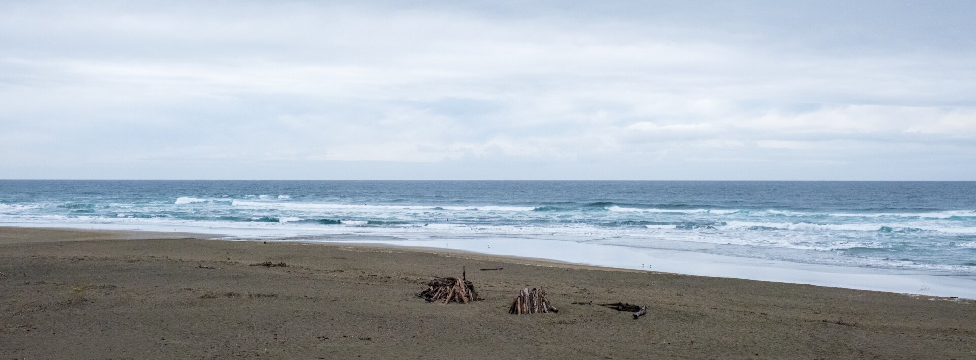 A beach with two driftwood huts on it, on a clear but cloudy day. [Fuji X-T5 / Fuji 23 1.4]