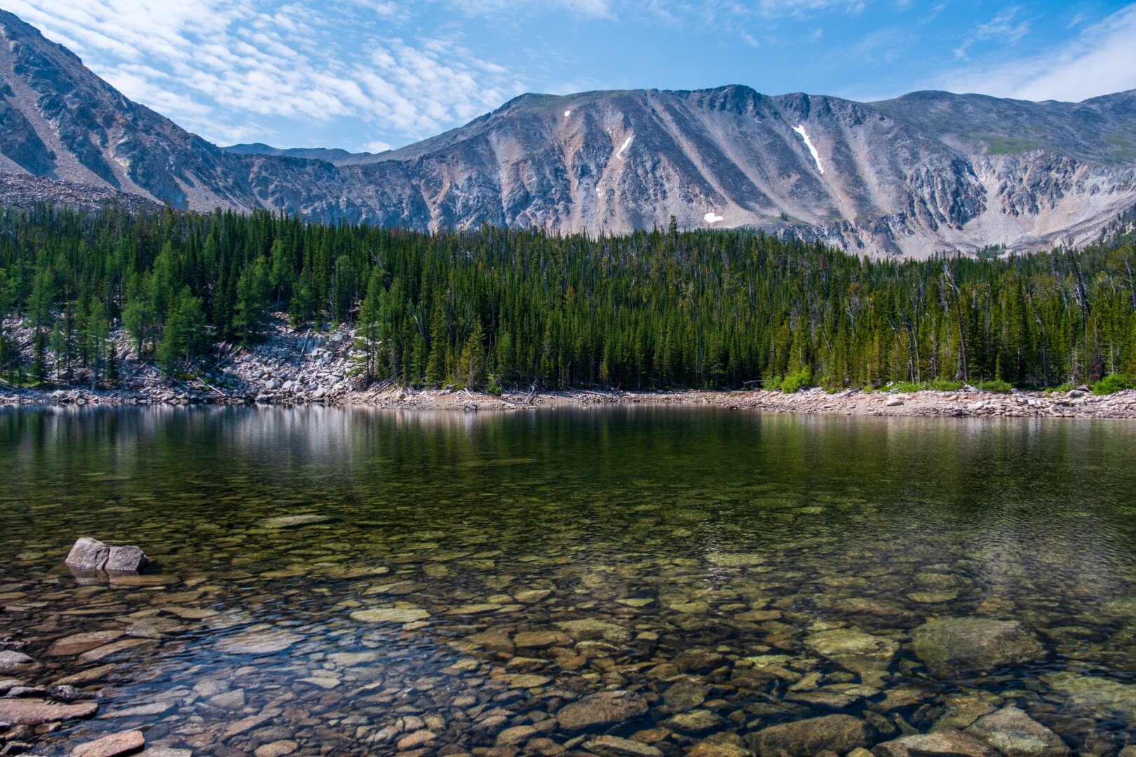 Many rocks lie under the surface of a lake, with mountains and forest behind. [Fuji X-T5 / Tamron 18-300]