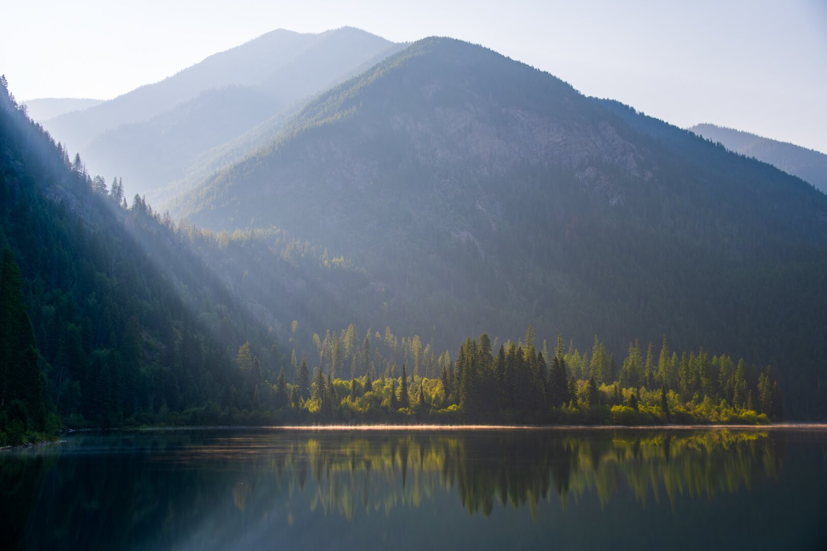 The sun shines on a lake through mountains, lighting up a tree-covered low spot which reflects on the lake. [Fuji X-T5 / Tamron 18-300]