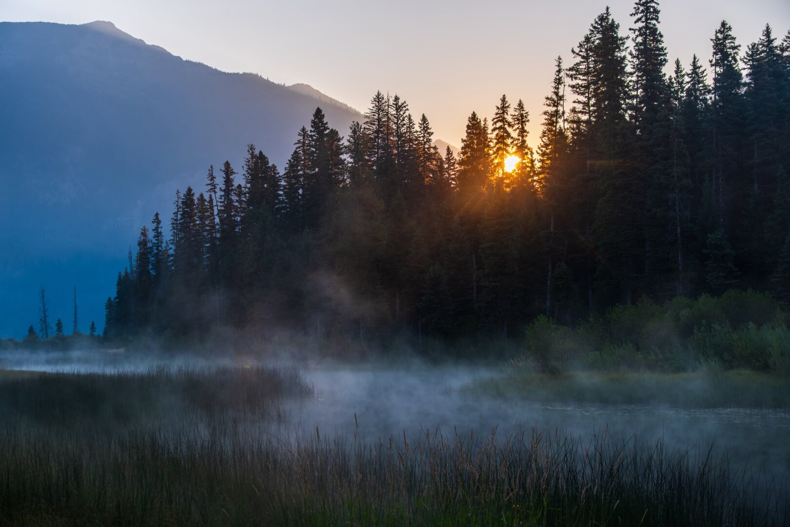 The sun rises over a misty little river feeding a lake, with mountains behind. [Fuji X-T5 / Tamron 18-300]