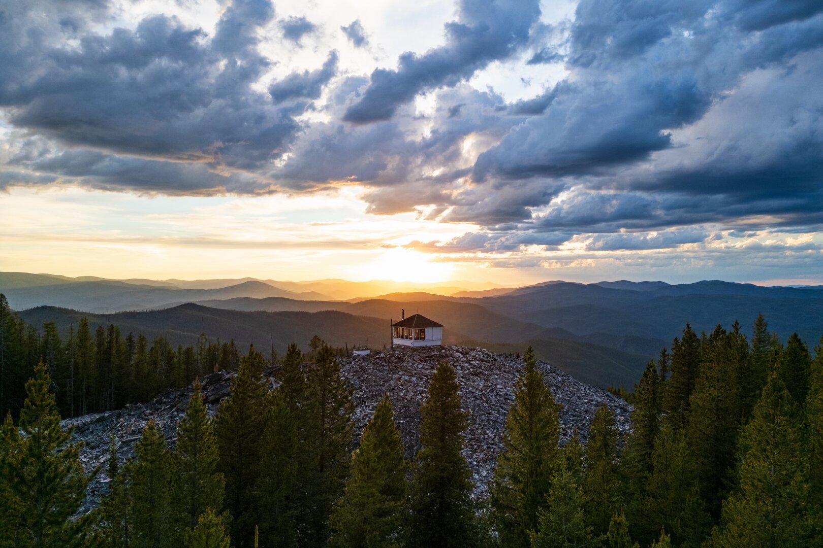 A fire lookout stands on a rocky hill amidst trees, with mountains and clouds behind. [DJI Mini 3 Pro]