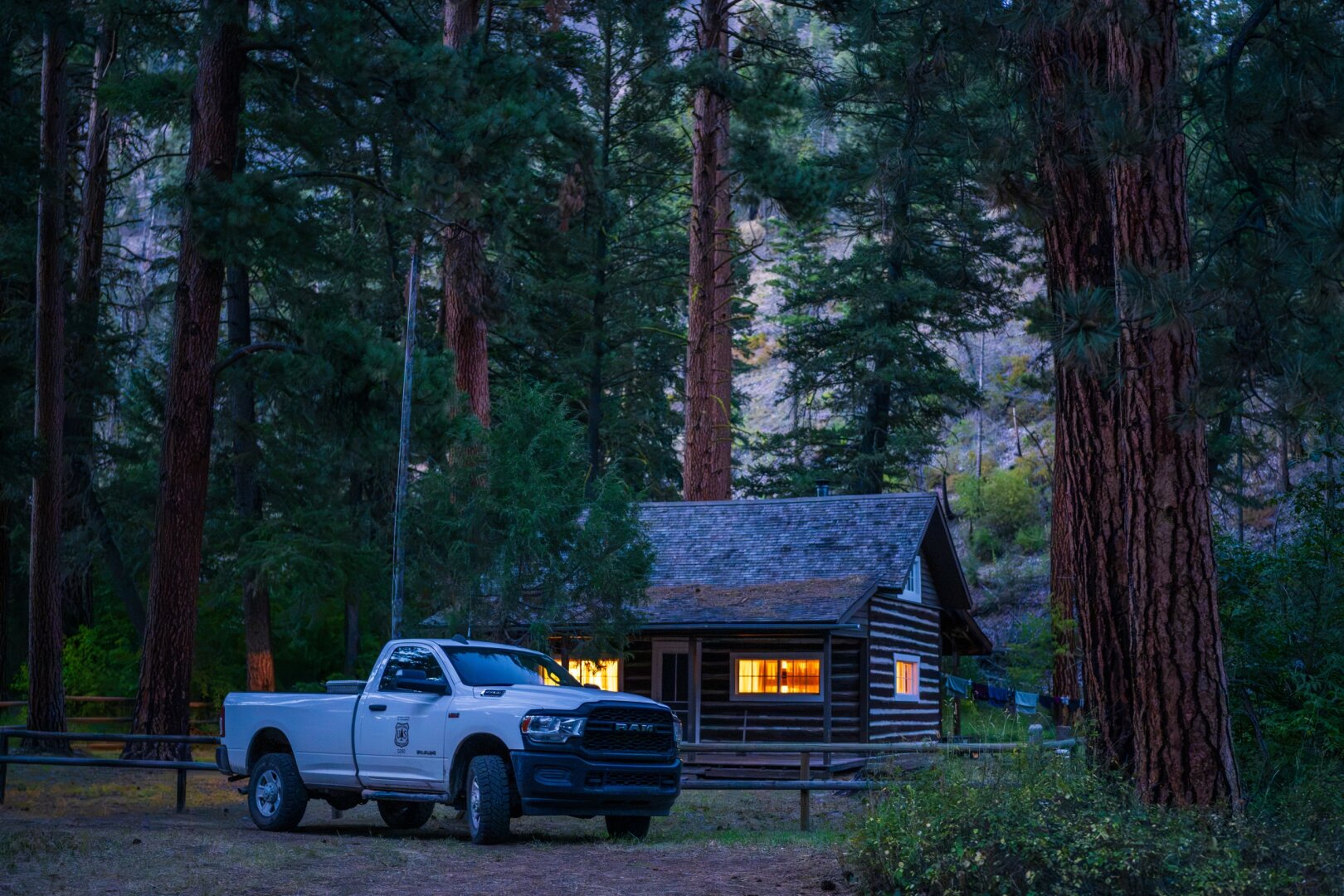 A white truck with Forest Service badging sits in front of a log cabin at dusk with its lights on. [Sony A7R V / Sirui Aurora 35]