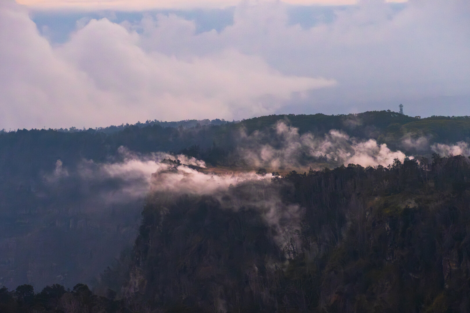 A far-away cliffside is covered in trees; steam rises and is blown off the edge. [Fuji X-T5 / Tamron 18-300]