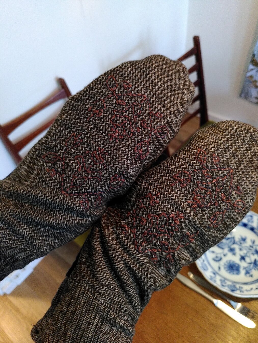 Photo of two hands in mittens made of a fine brown trouser fabric with stylized flowers embroidered on the backs in brown thread.