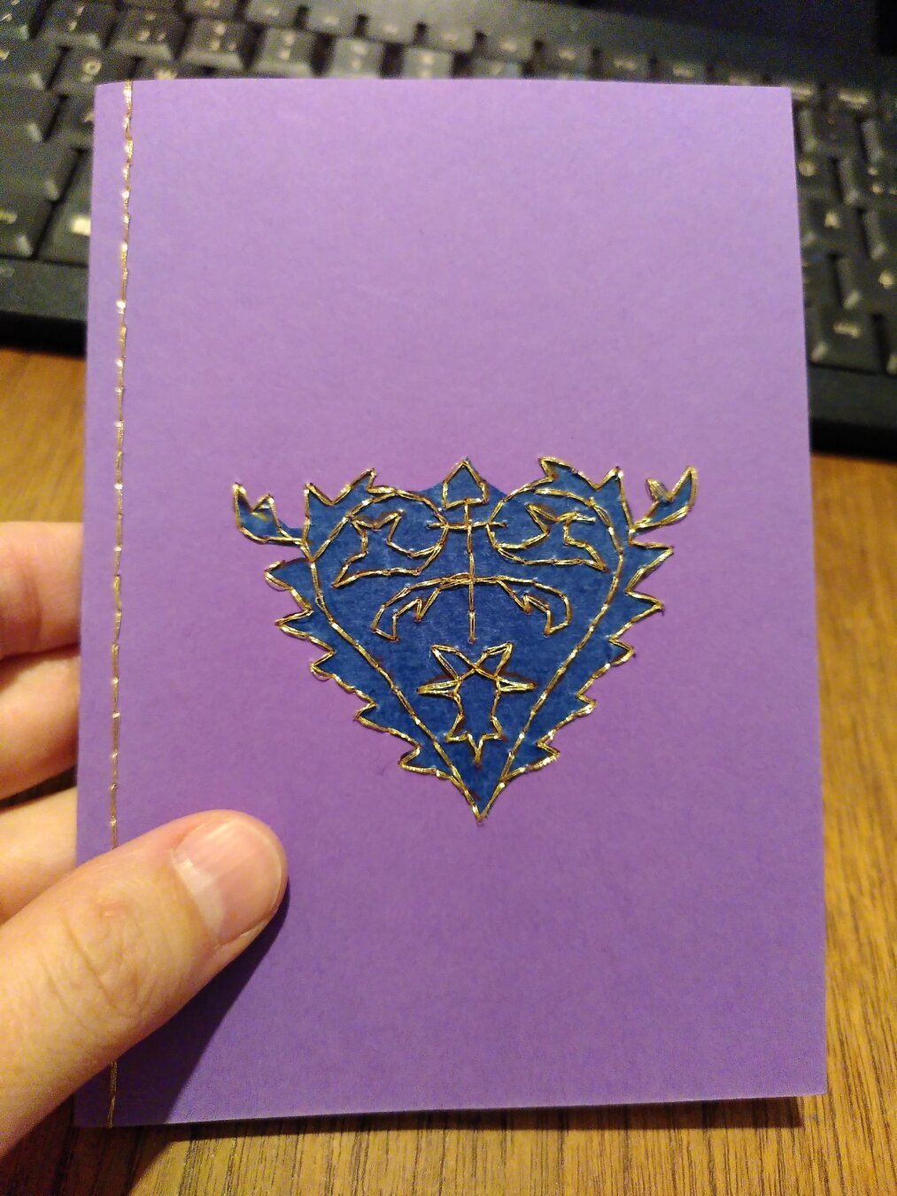 Purple card with an elaborate heart pattern embroidered in gold thread, with the heart itself filled in with dark blue transparent paper.