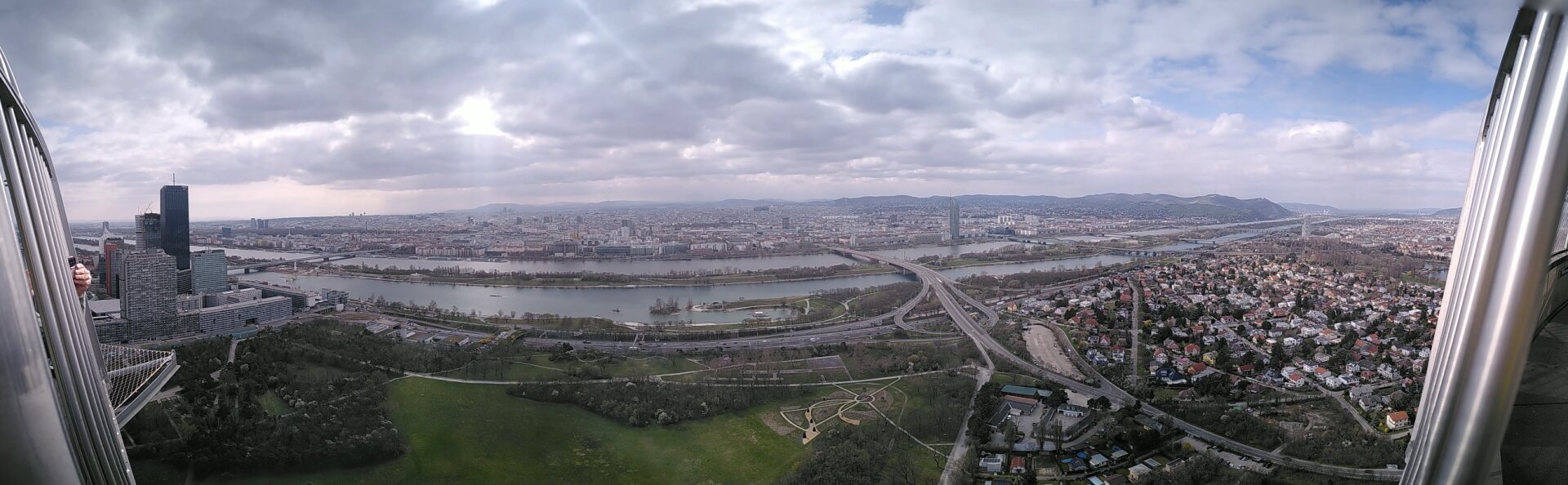 Man sieht eine breite Panoramaansicht  der Stadt Wien vom hohen Donauturm aus. Ganz vorne noch grün, dann neue Donau, Donauinsel und Donau. Dahinter die Stadt. Bewölkter Himmel mit ein paar Sonnenspitzen. Blickrichtung Südwest