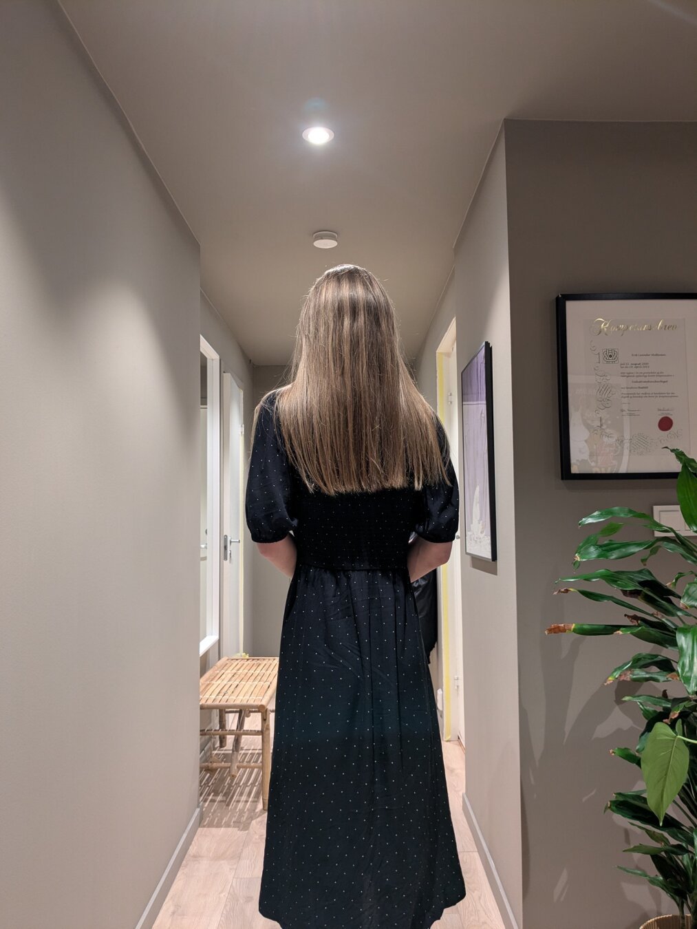 In a quiet hallway, a young woman stands alone. 

A simple wooden bench sits discreetly to the side, while a potted plant adds a touch of sterile life to the space. 

The scene is lit by unremarkable ceiling fixtures.