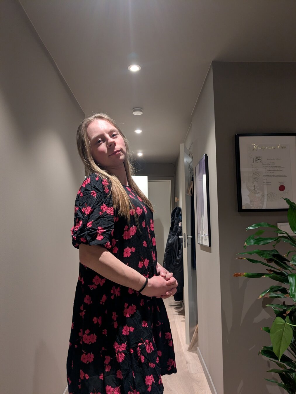 A woman stands in a muted hallway of her apartment.

A framed certificate and a large potted plant occupy the space on the right, adding small details.