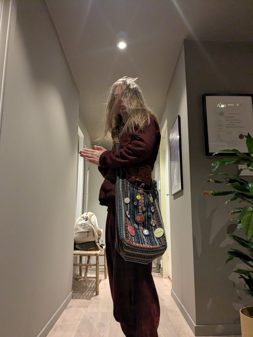 In a hallway of an apartment a man stands quietly.

A backpack rests on a wooden stool in the background, while a certificate frame and a potted plant add to the domestic setting.

The lighting is subdued, casting shadows that accentuate the stillness of the scene.