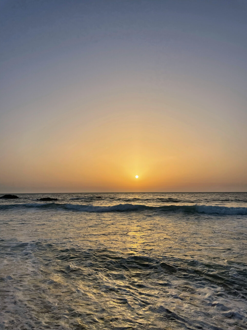 Sunset over the ocean. The sky is hazy but without clouds. The ocean is filled with small wave ripples, giving it lots of texture. There are small waves at the distance