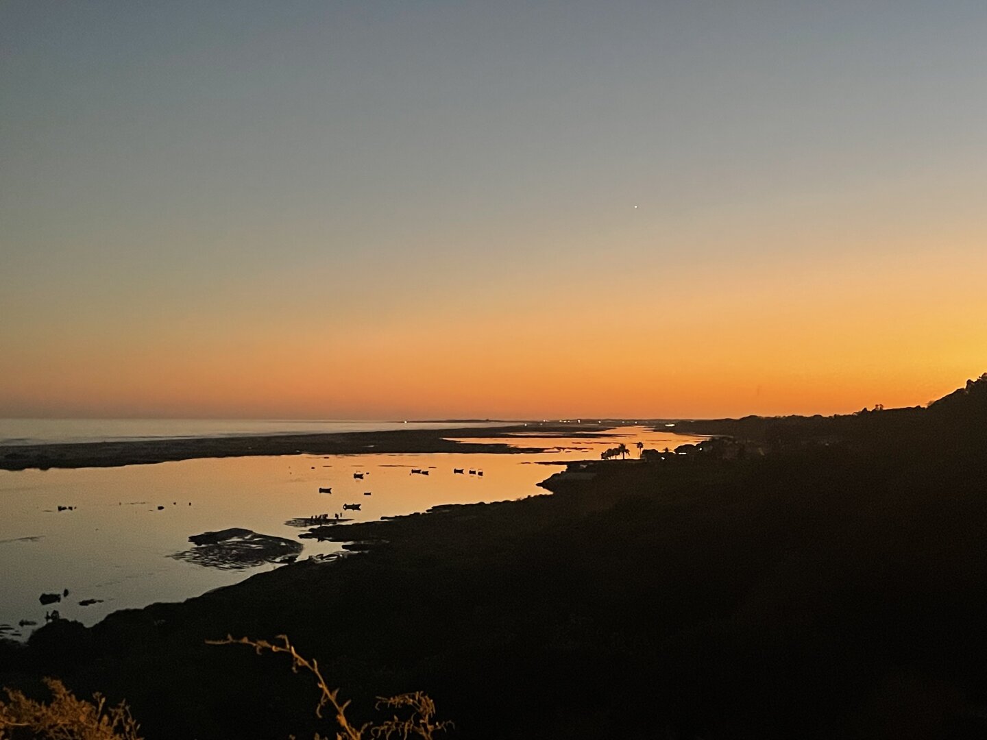 Sunset over a beach. The photo shows the last orange tones of a sunset. On the distance there’s a couple of palm trees and some boats on the water