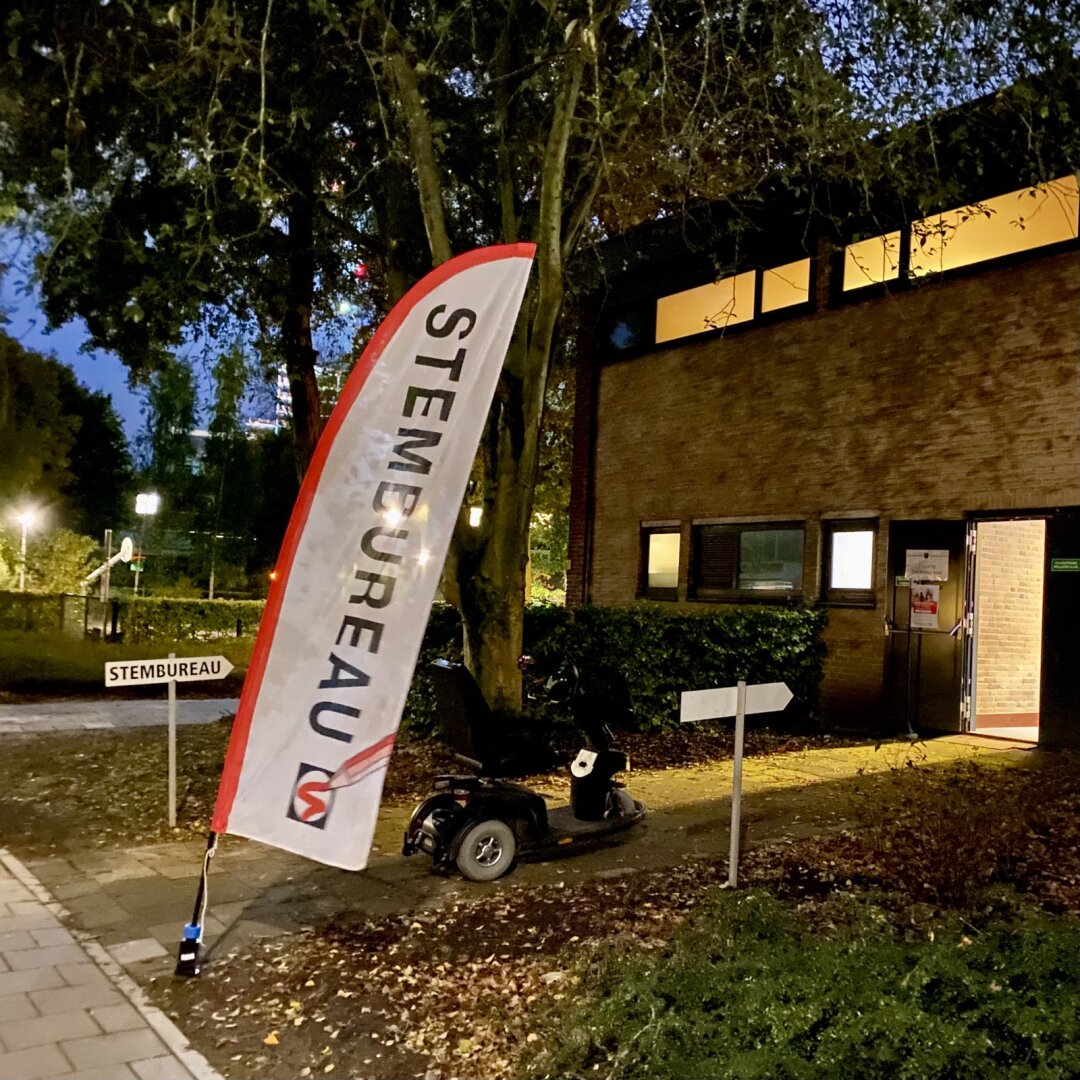 Dutch voting office in the night, with flags and signs saying “stembureau” / voting bureau. Signs lead to  a welcoming opened door with lights shining