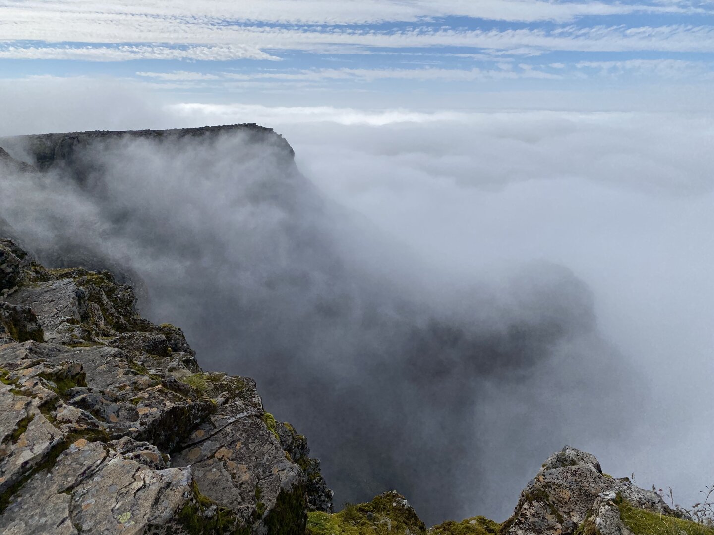 Picture taken from Ben Nevis summit, with a blue sky, and clouds below in the valley.