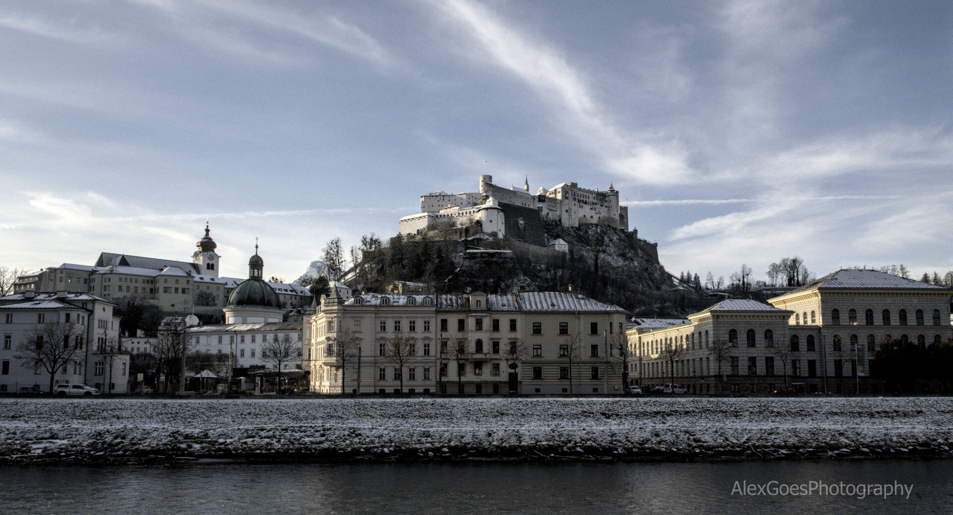 View of an old european city. A river flows in the foreground from left to right and behind it the city raises up to a forstress on a hill towering above. The city is dusted with a light coat of snow. The sky is grey/light blue with soft snow.