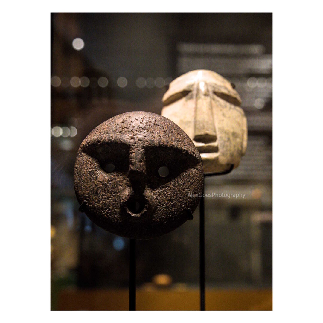 Two Pre-Columbian America stones shaped like two faces. On the left in foreground, a round dark brown/black one with strong eyebrows, perfrectly round small holes for eyes and a small o mouth. Behind, on the right, a white one more oval, with eyebrows sloping to the sides, long nose and straight mouth. They lights come from above, making shadows below the strong eyebrows of the round face in foreground.
