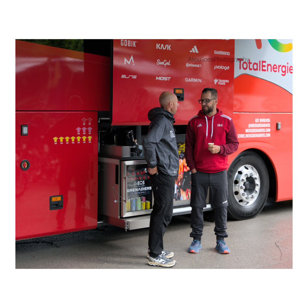 Two members of the Ineos Grenadiers pro cycling team having a chat in front of their bus and their little fridge with on top a small coffee machine coming out of the bus. On the left, on the bus panel, the yellow, pink and red jerseys counting the times the team won the three grand tours. The panel above the coffee machine and behind the two people has various sponsors arrayed in a grid. The man on the left wears black pants and grey hoodie while the one on the right, holding a tiny coffee cup has black pants and red hoodie.