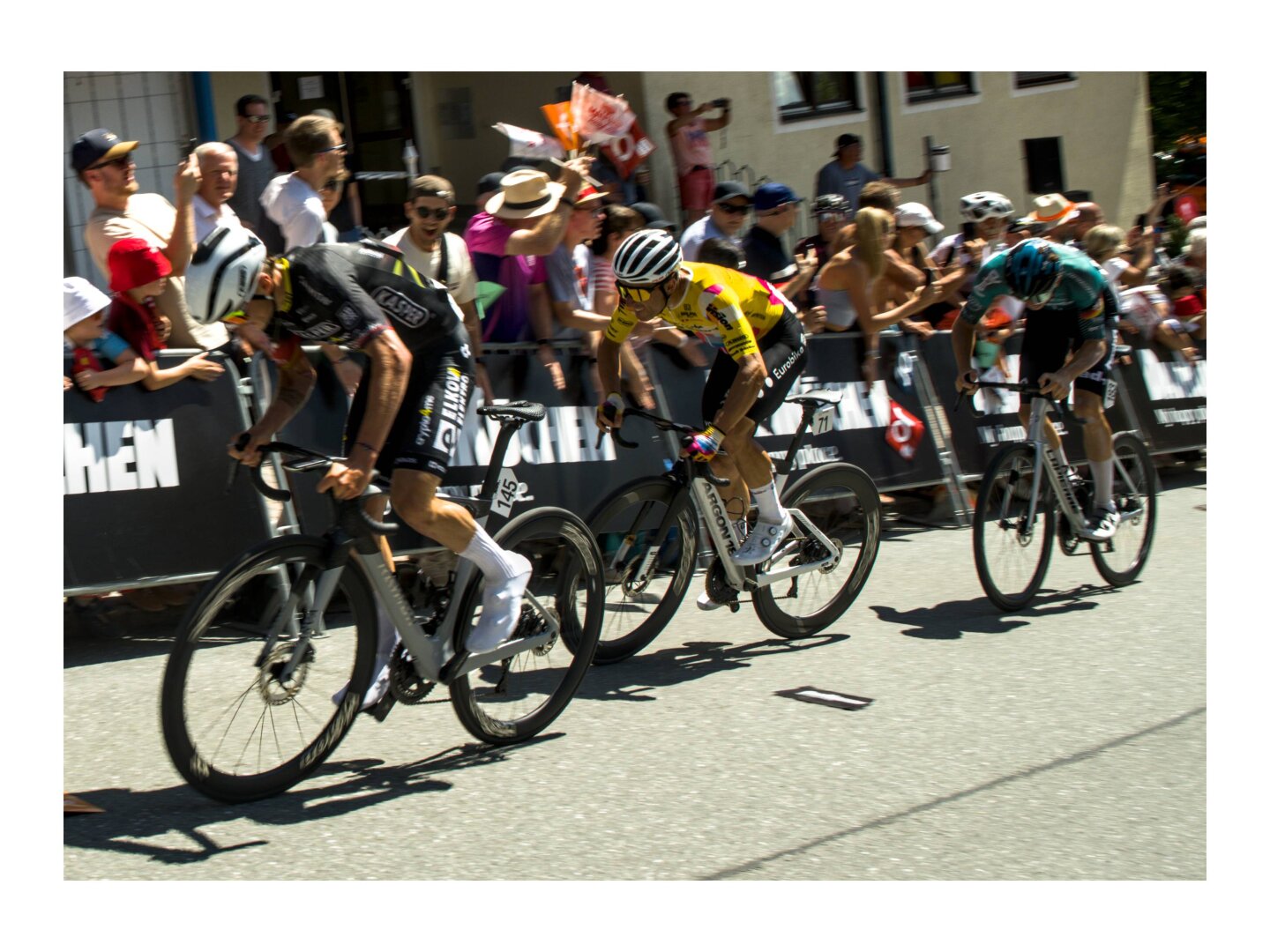 3 riders from 3 different teams give it all on the climbing roads of the Tour of Austria. Behind them, the public at the barriers, slightly blurred and cheering under the bright sun.
