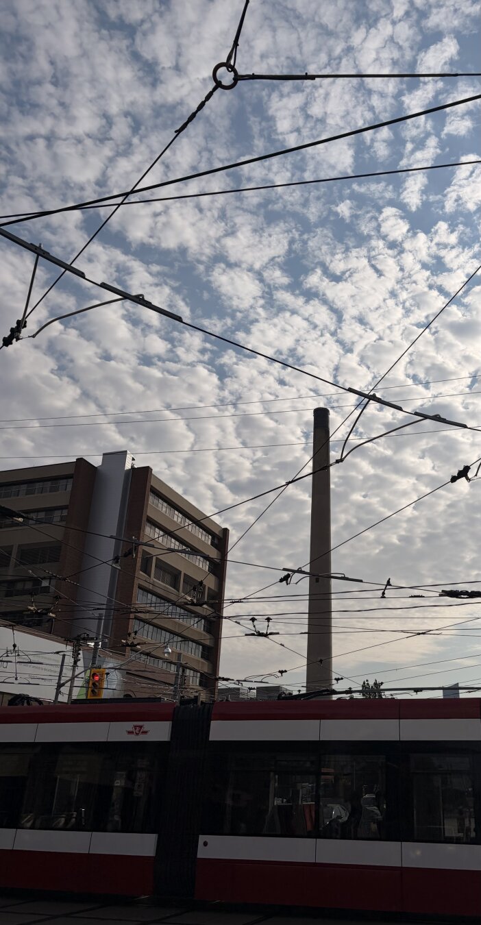 Upward angle photo of TTC streetcar, overhead wires, urban mid-rise (brown with linear windows), smokestack on a background of cottony clouds with a blue sky. Early morning slanting sunlight.