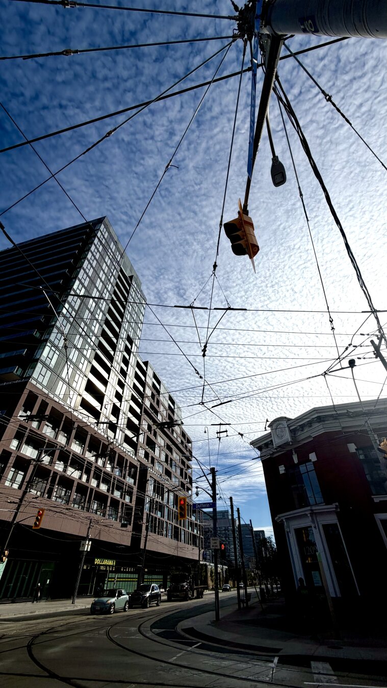 Upward angle photo showing an urban intersection with overhead wires, stoplight and high clouds. Mid-level multi-storey buildings are visible across the intersection.