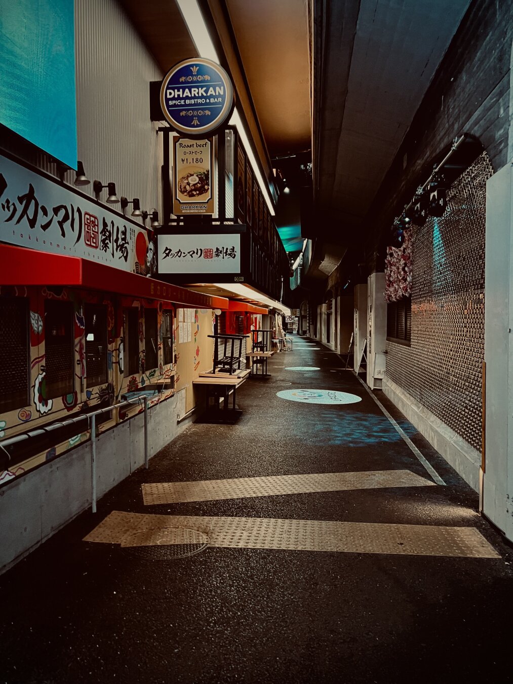Food shops tucked in underneath a rail bridge, with neon lighting and brick streets, reminiscent of an alley.  It’s empty at the moment of this photo.