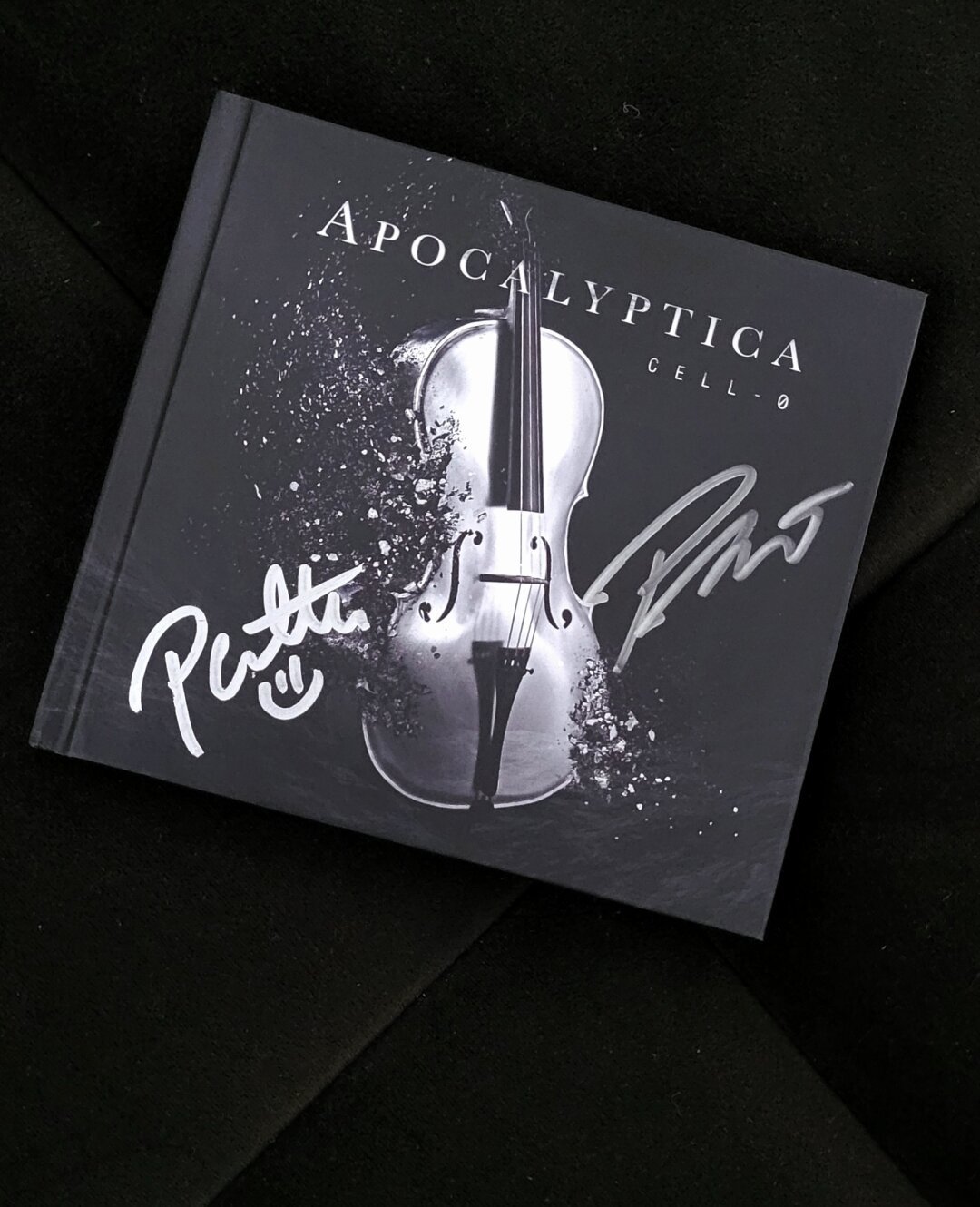 A CD album on a dark brown background. Album cover is anthracite, almost black, a silver cello leaning to the right is the central motive. The cello is disintegrating. Band name Apocalyptica with big silver characters under the upper edge, album name Cell-0 to the right with smaller characters under band name. Two signatures in silver marker are to the left and right of the cello)