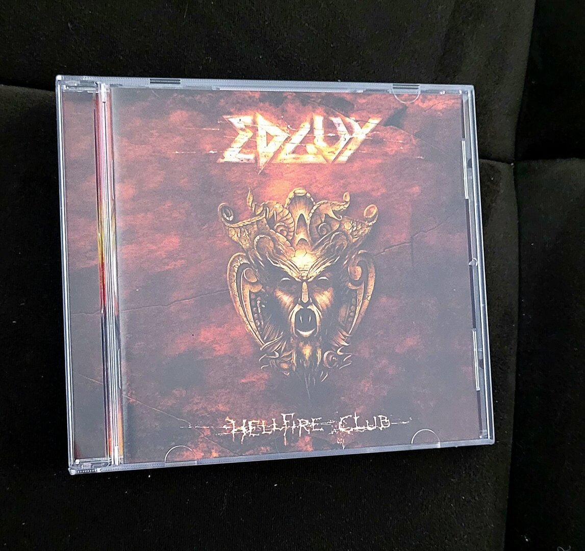 A picture of a CD on a dark brown background. CD in a plastic jewelcase, cover is dark red clouds with a big golden screaming mask of a creature resembling a lion and a bear at the same time. Artist name Edguy with big burning golden letters under the upper edge, album name Hellfire Club with scratchy but readable letters under the mask.