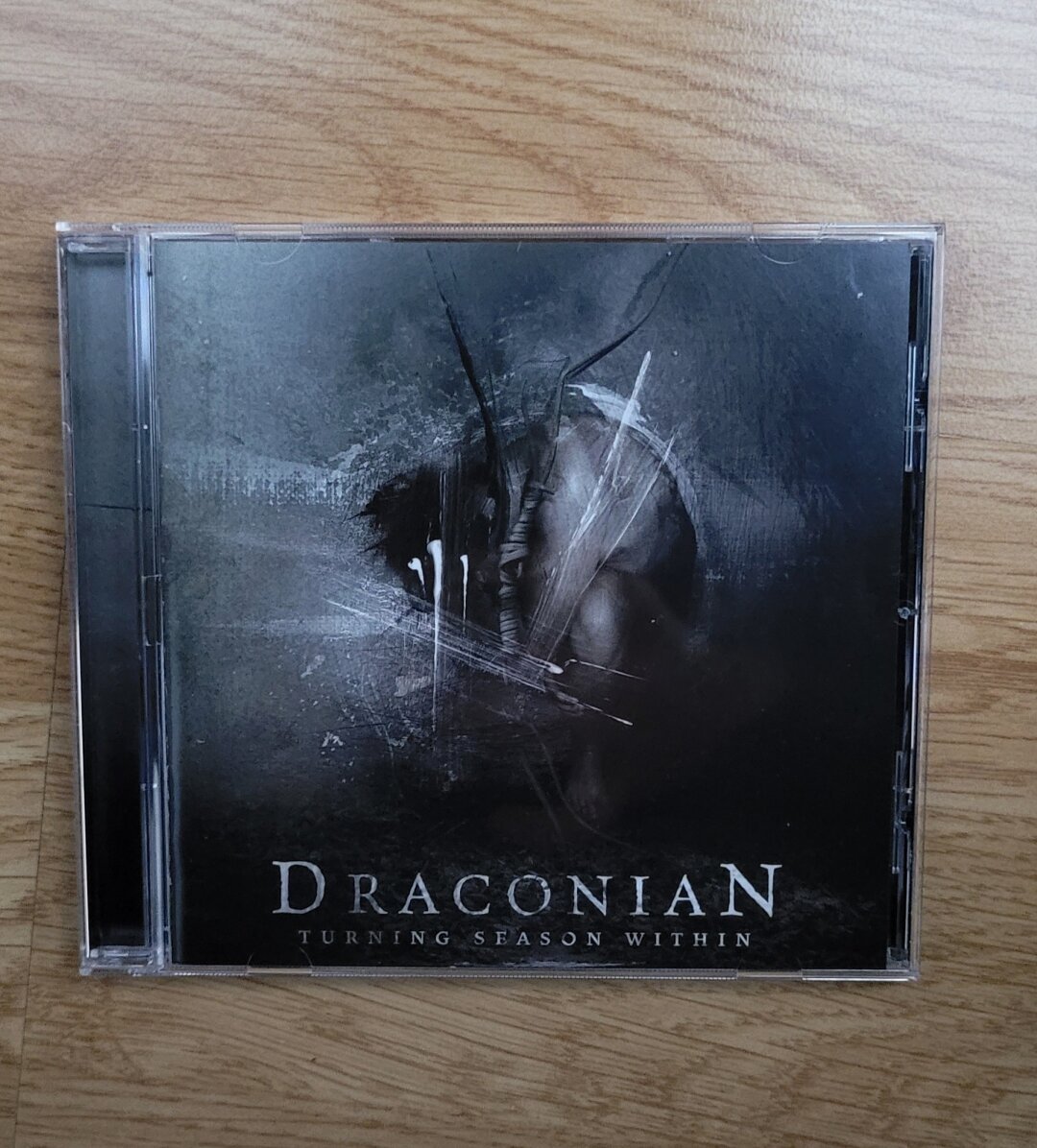 A CD album on orange-brown flooring. Album cover is dark grey green, an impression of a person huddled together is in the middle, over the impression there are a few brush strokes of white paint. Band name Draconian above the lower edge in big characters, album name Turning Season Within with smaller characters underneath.