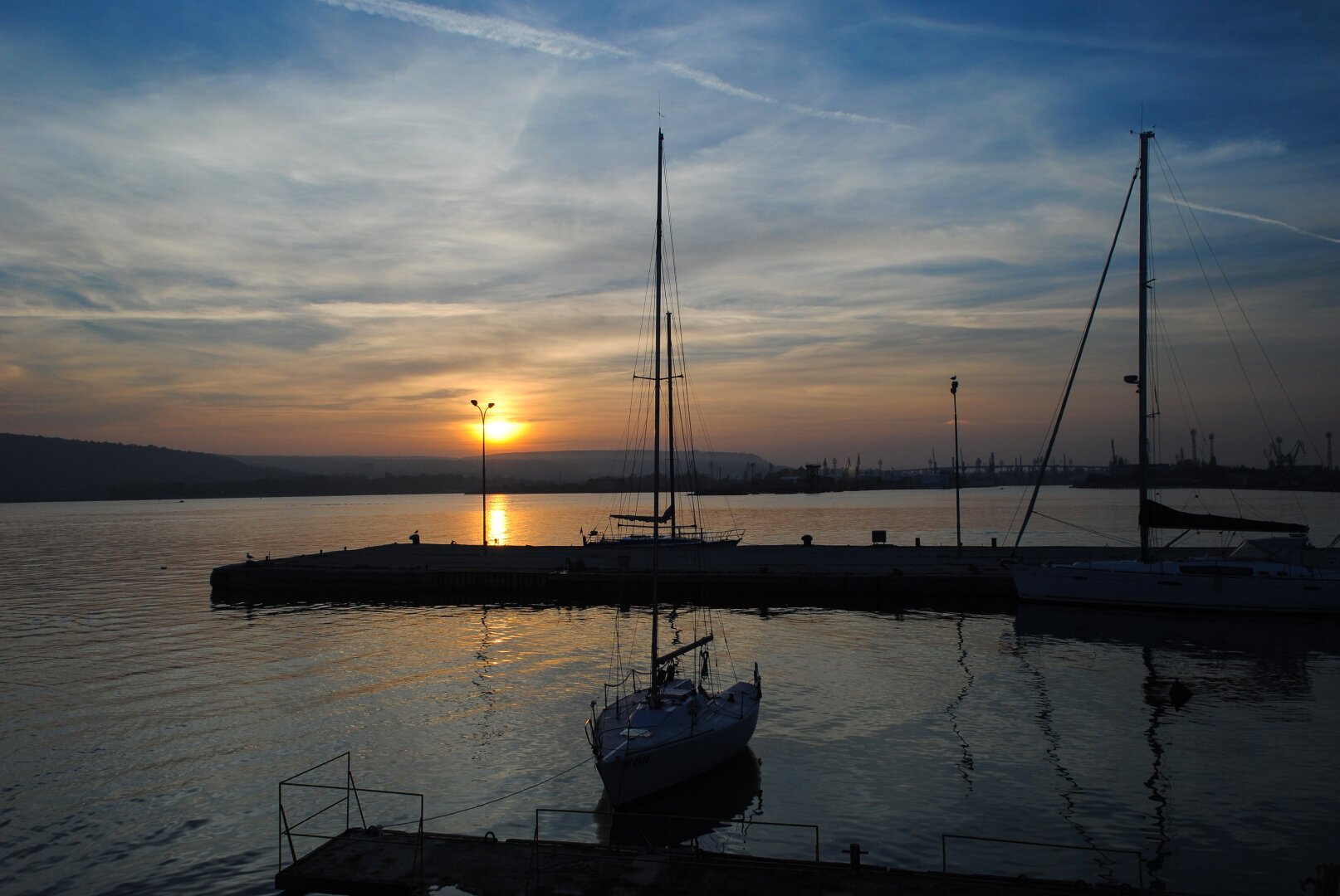 A sea view at sunset, with fishing boats and piers in the foreground and multiple ship masts in the background