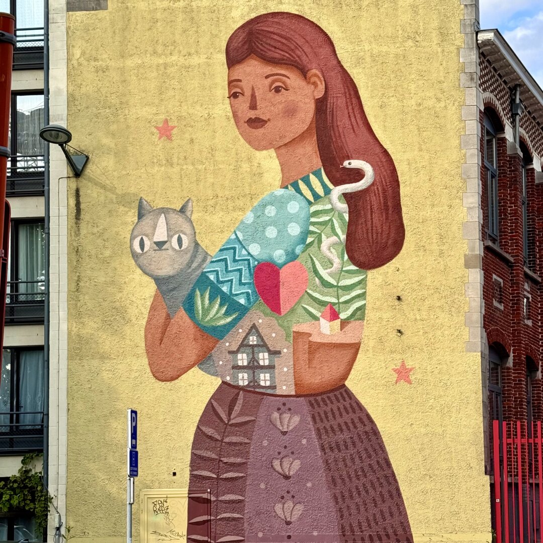 A large, colorful mural is painted on the side of a yellow building. It shows a woman with long, reddish-brown hair holding a grey cat in her arms. A white snake wraps around her hair and shoulder. Her clothes are decorated with different symbols: a red and pink heart, green leaves, a small house, a red star, and a paper boat. Her skirt is purple with flower patterns. The woman’s expression is calm and kind. The background of the wall is plain yellow with two red stars. On the left side, part of a modern building with balconies is visible, and on the right, there is a red brick building. A streetlamp and a blue parking sign are also in the scene. The artwork feels peaceful and warm, showing a connection to nature and home.