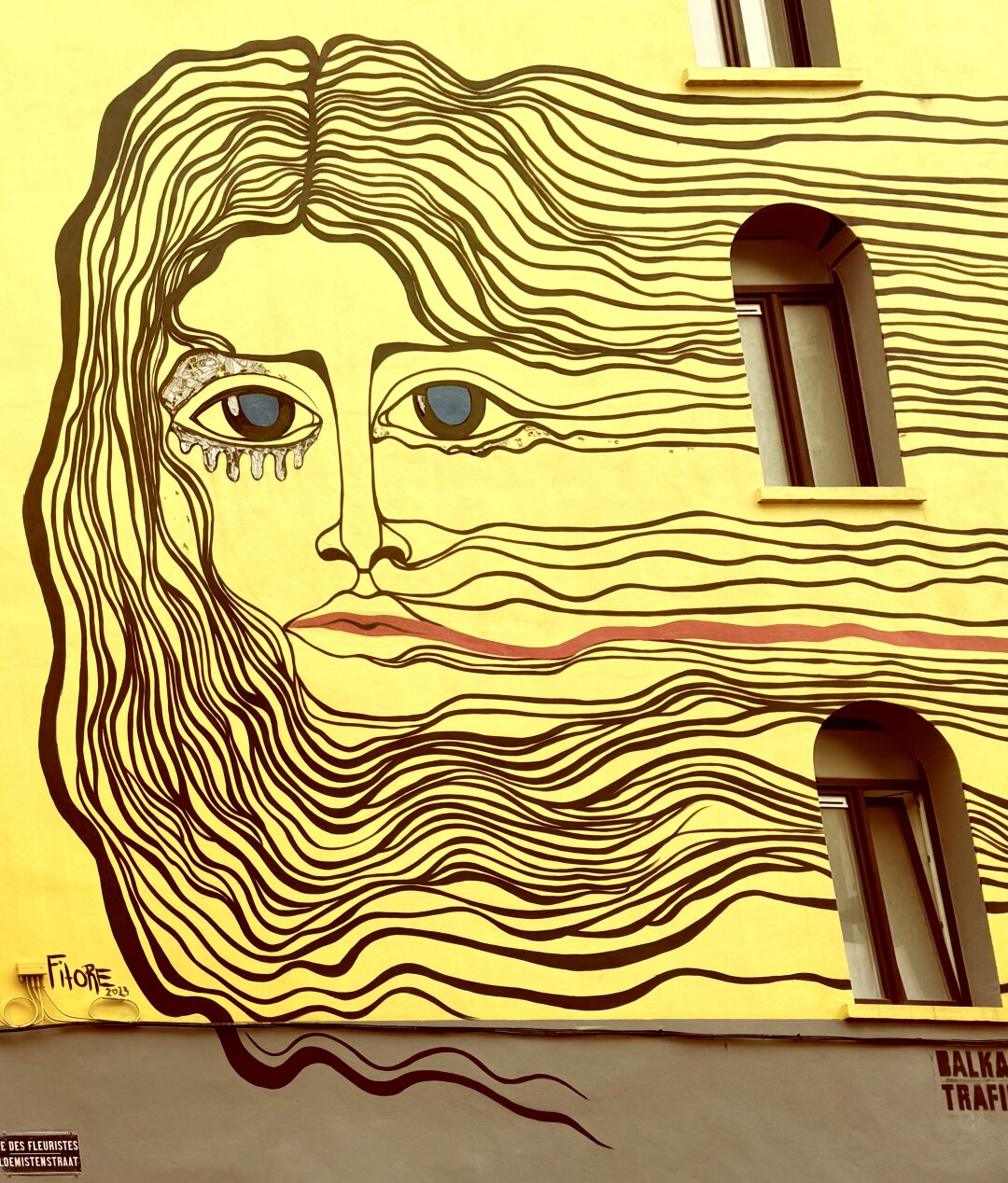 This image shows a large mural painted on the side of a yellow building. The artwork is a stylised face of a woman covering several floors. Her long hair is made with many flowing dark lines that stretch across the wall and around the windows. Two windows are placed where her eyes would be, and another window sits in the area of her hair. One eye is decorated with white, melting shapes that look like tears. Her lips are simple and painted in light red. The face has a calm but mysterious expression. At the bottom left, the artist’s signature “Fitore 2023” appears. The wall colour is bright yellow, which makes the dark lines stand out strongly.