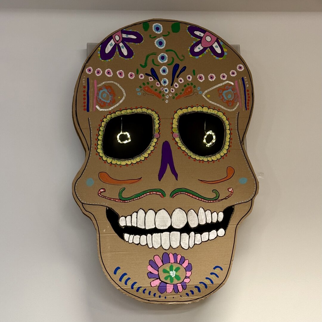 Un adorno en forma de calavera está colgado en una pared blanca. Es una calavera de estilo “calavera mexicana” hecha de cartón marrón y decorada con colores vivos. Los ojos son grandes círculos negros con luces reflejadas. Alrededor de los ojos hay un borde amarillo con pequeños puntos verdes. La calavera tiene dibujos florales y líneas curvas en colores como rosa, verde, azul, morado y naranja. En la frente hay flores y puntos decorativos. La nariz es una forma sencilla en color morado. La boca está abierta y muestra muchos dientes blancos bien marcados. En la barbilla hay una flor rosa y verde rodeada de puntos azules. El estilo es alegre y festivo.