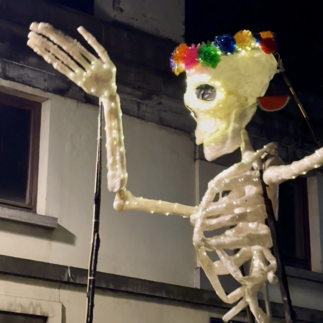 This image shows a large, decorative skeleton standing outside at night. The skeleton is not just white bones—it is brightly lit with small, warm lights along its arms, hands, and body, making it glow in the dark. The skeleton’s head is unique: it wears a colorful headdress made of big, fluffy flowers in many colors like pink, yellow, green, and blue. The skeleton is posed with one arm raised, as if it is waving or greeting someone. It stands in front of a building, and you can see part of the building’s roof and a window behind it. The scene looks festive and creative, likely part of a celebration or holiday decoration. The lights and colors make the skeleton look cheerful and eye-catching.