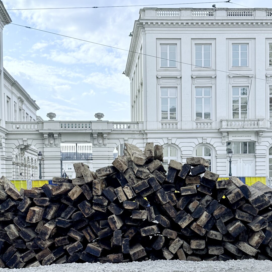 A large pile of dark, scorched wooden beams lies on rough gravel in the foreground. Behind it stands a bright white, neoclassical building with tall rectangular windows, heavy cornices, and a small sign by the door that reads “Musée Magritte Museum.” A stone bridge-like passage with a balustrade connects two wings of the building, and an iron gate is visible through it. Yellow-and-blue construction barriers line the site, showing that road or track work is in progress. Thin tram or power lines cut across a pale blue sky with soft clouds. The contrast between the dirty, broken wood and the clean, elegant facade creates a strong visual tension.