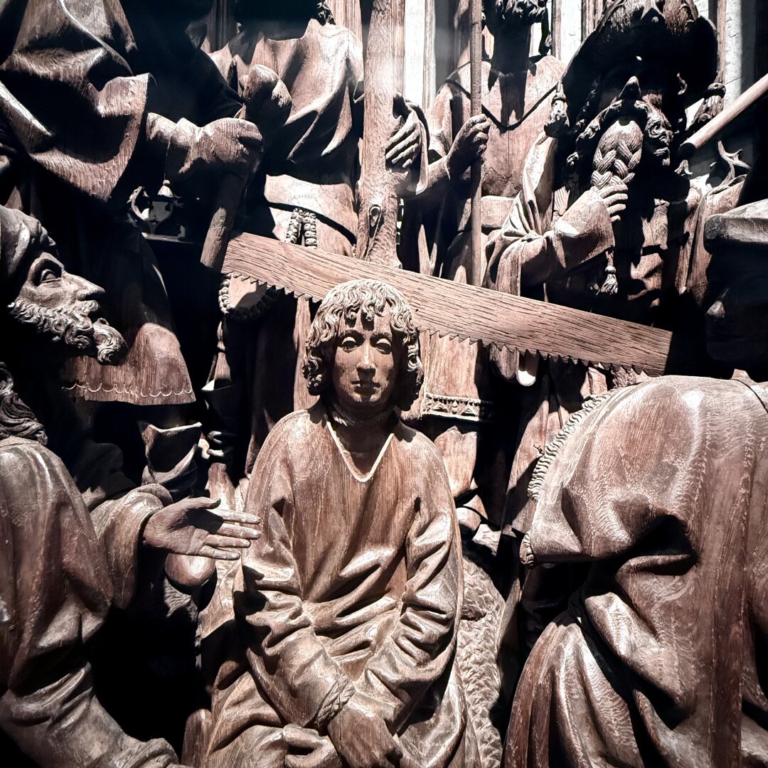 This is a detailed wooden carving showing many people in robes. In the center, a young man with curly hair sits with his hands crossed on his lap, looking forward calmly. Around him, other figures stand and talk, with some holding objects like a saw and sticks. The carving shows careful details in the hair, faces, and folds of the clothes. The wood is a warm brown color, and the light highlights the smooth surfaces and fine lines, making the scene look alive and full of movement.