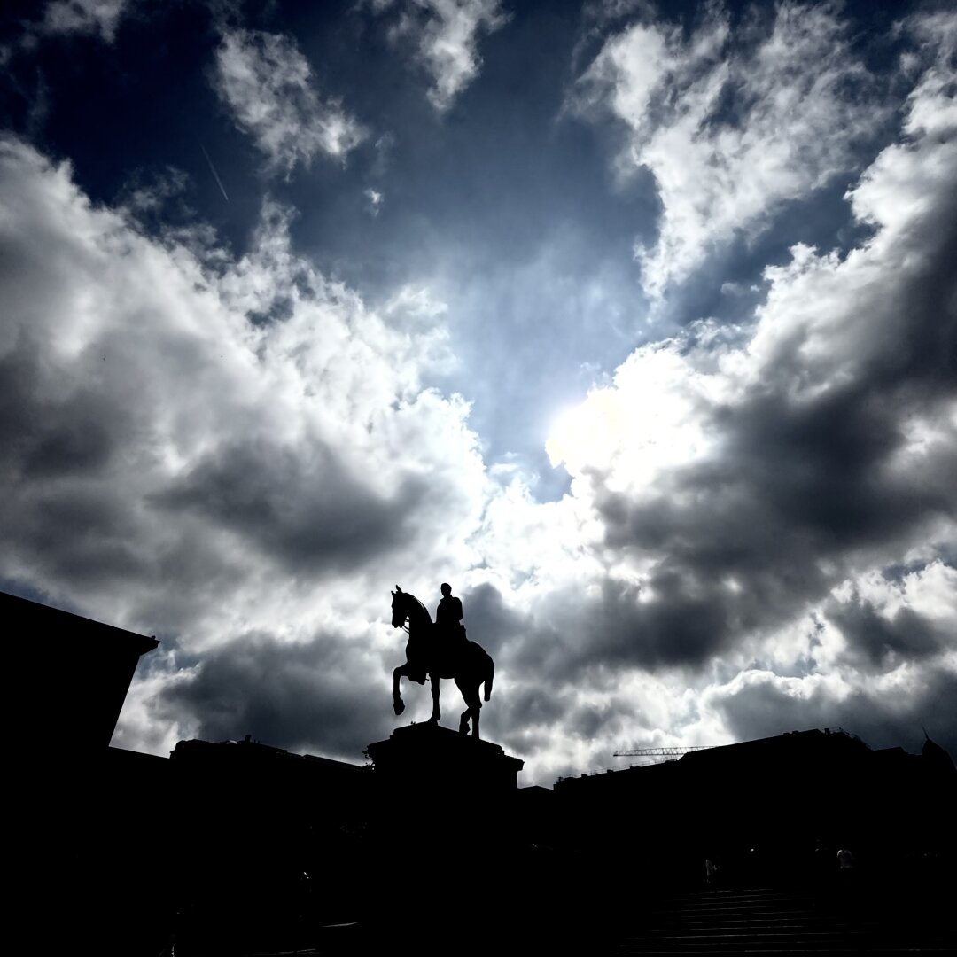 A silhouette of a large statue shows a man riding a horse. The statue stands on a tall base in the center of the image. The horse has one front leg raised. The sky behind the statue is full of dramatic, dark clouds. The sun is hidden behind the clouds, but bright light shines through, creating a strong contrast between the sky and the dark shape of the statue. The buildings around the statue are also in shadow and hard to see clearly. The mood of the image is powerful and dramatic because of the strong light, dark shadows, and the cloudy sky.