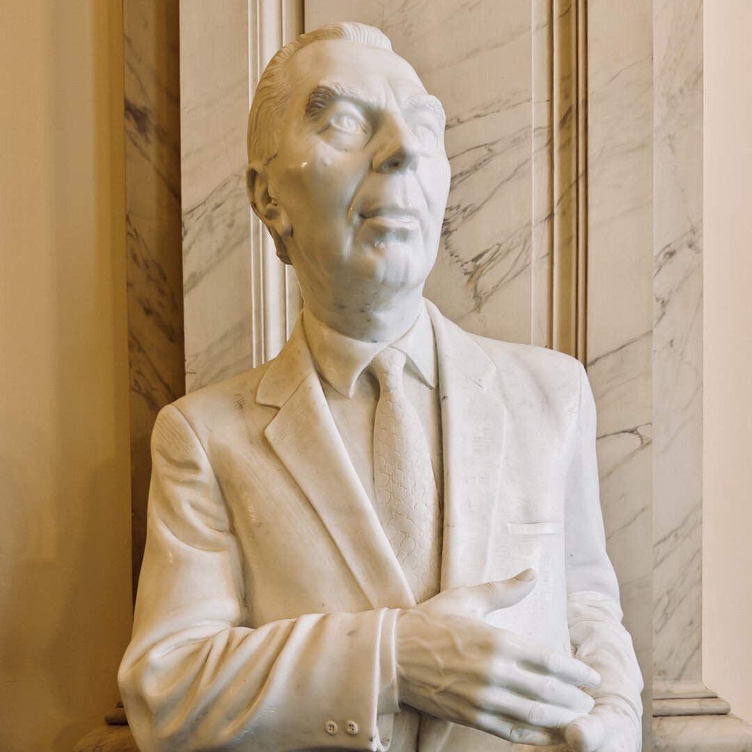 A white marble statue of a man standing in front of a polished marble wall. The man is wearing a formal suit with a tie and a collared shirt. His jacket has a pocket on the left side and buttons on the cuffs. One hand is placed over his chest, with the fingers gently resting on the jacket, while the other arm is relaxed. His face looks calm and thoughtful, with short, combed-back hair and detailed facial features such as a pointed nose, full lips, and deep-set eyes looking slightly upward. The texture of the tie shows a fine pattern. The background is made of smooth, light-colored marble with dark gray veins. The statue gives a sense of dignity, respect, and calm leadership.