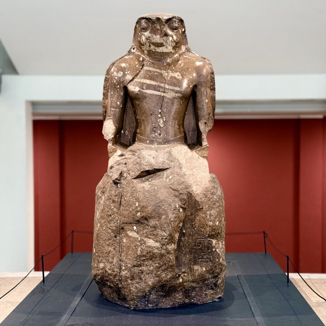 A large stone statue stands on a black platform in a museum with red walls. The statue shows a figure with a human body and a head shaped like a frog or toad. The chest is detailed with a necklace, while the arms are broken at the elbows. The lower part of the statue is rough and unfinished, showing natural rock texture. The surface has many white spots and cracks from age. The figure’s big round eyes and flat mouth give it a strong, calm look. A doorway is visible behind the statue.