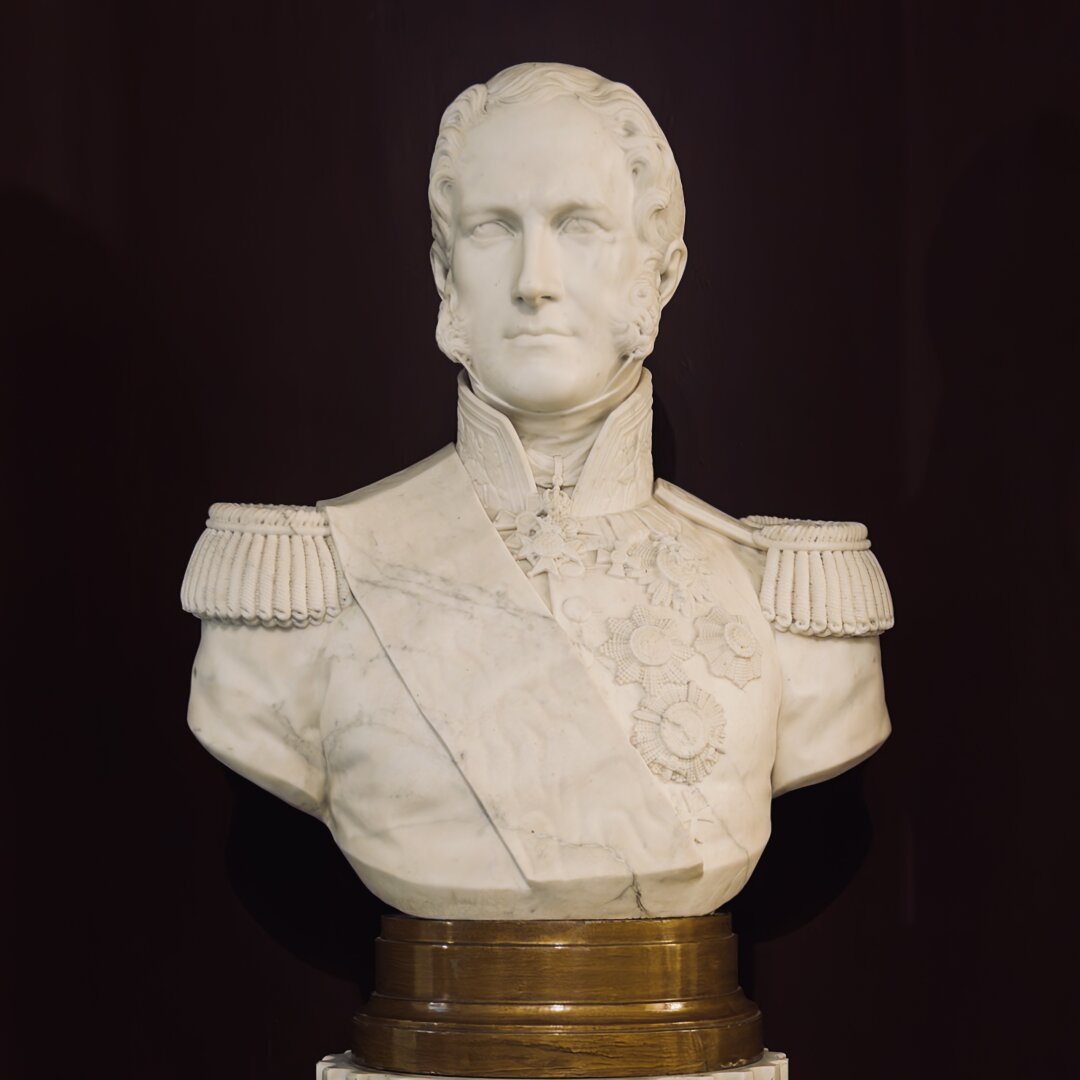 A marble bust of a man is shown against a dark background. The man has short, wavy hair and strong facial features, including high cheekbones and a straight nose. He wears a decorated military uniform with detailed shoulder epaulettes and a sash across his chest. Several medals and star-shaped badges are carved on his chest, showing his high rank. The collar is tall and stiff, and the uniform is carefully sculpted to look realistic. The bust is placed on a round wooden base. The sculpture has a smooth, white surface with fine lines to show texture in the clothing and hair. The man’s face looks serious and proud, facing forward with a calm expression.