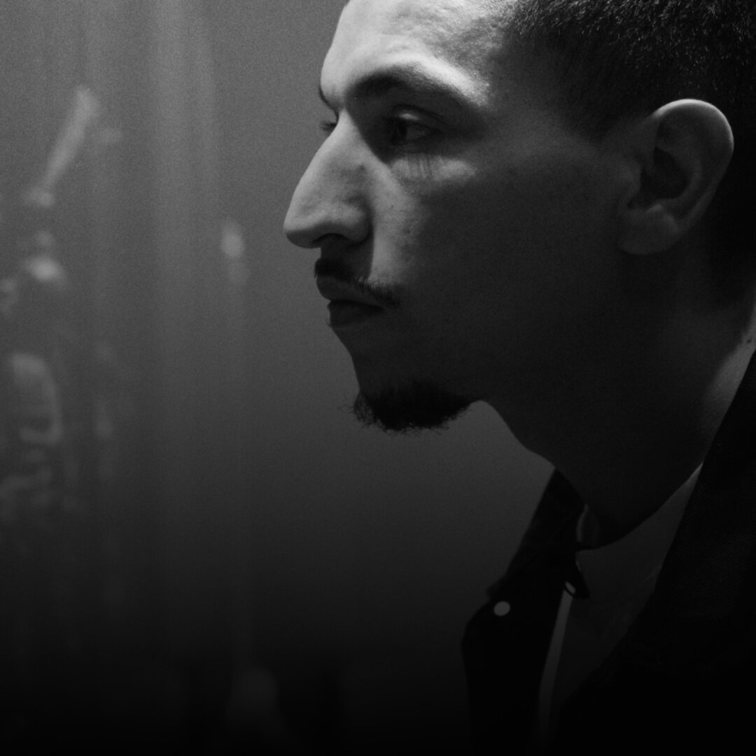 A black and white photo shows a man in profile, facing left. He has short dark hair and a trimmed goatee. His nose is long, and his lips are closed, giving him a calm, serious look. He wears a light shirt under a dark jacket with a visible button. The background is blurry and dark, with a faint shape that could be a microphone stand, suggesting he might be in a recording studio. The lighting creates soft shadows on his face, showing his focused expression.