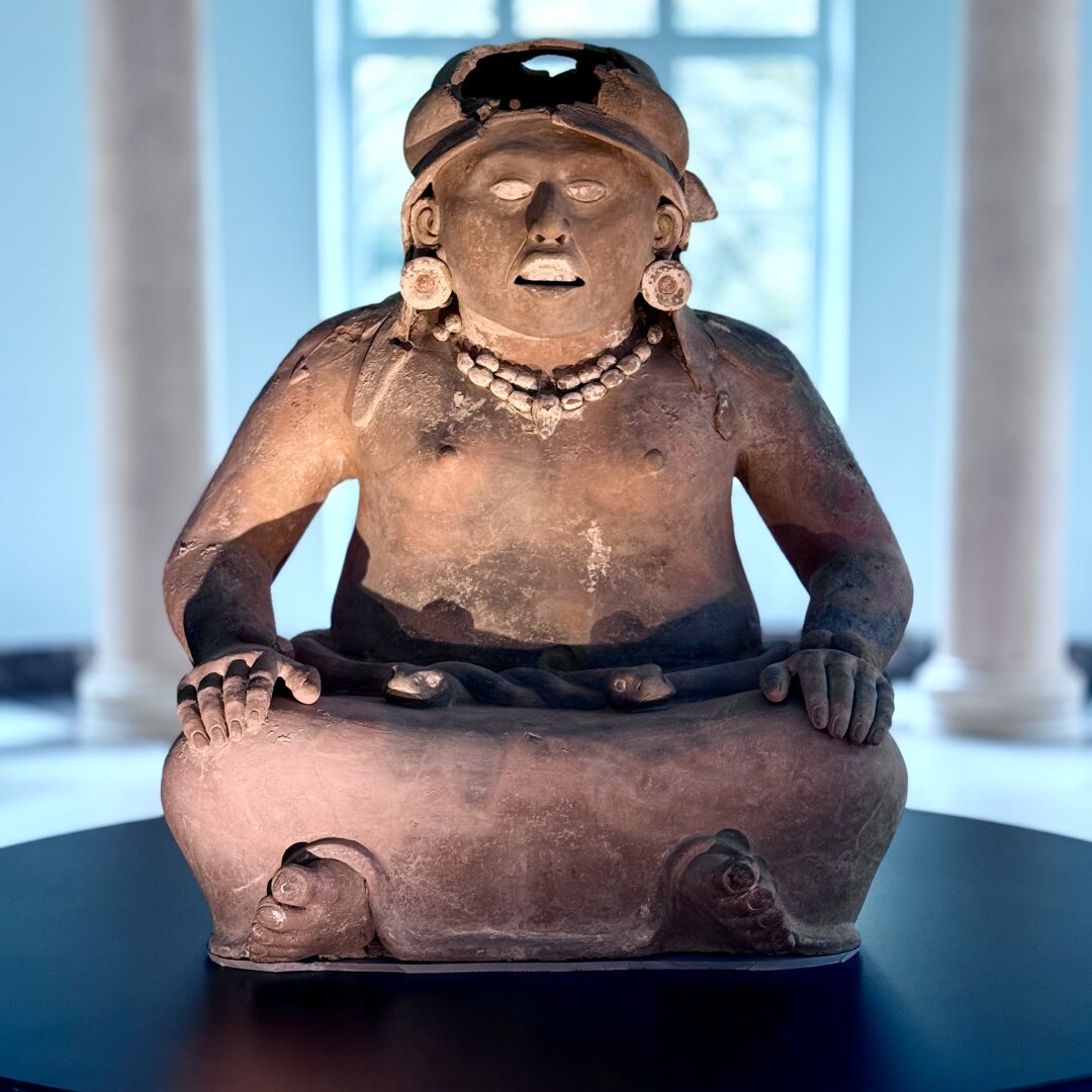 A clay statue shows a seated figure with a strong body, resting its hands on its knees. The figure wears a necklace with round beads and large circular earrings. It has a headpiece with a broken top and long ear decorations hanging down. The eyes are open and round, and the mouth is slightly open, showing teeth. The feet and toes are visible under the rounded base. The statue has a light brown color and sits on a black round stand in a bright room with tall columns and a large window in the background.