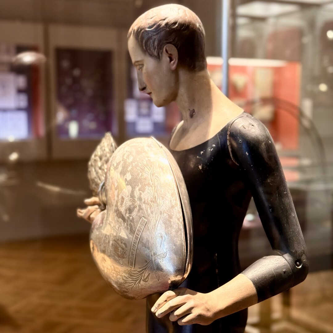 A wooden statue of a man is shown from the side, facing left. He has short hair and a calm face, looking down at a large, shiny silver shell he holds against his chest. The shell has engraved floral patterns and shines under the light. The man wears a tight, black outfit with visible small metal joints at his shoulders and elbows. The background is a museum room with glass cases, red walls, and blurry lights, creating a warm and quiet atmosphere around the statue.