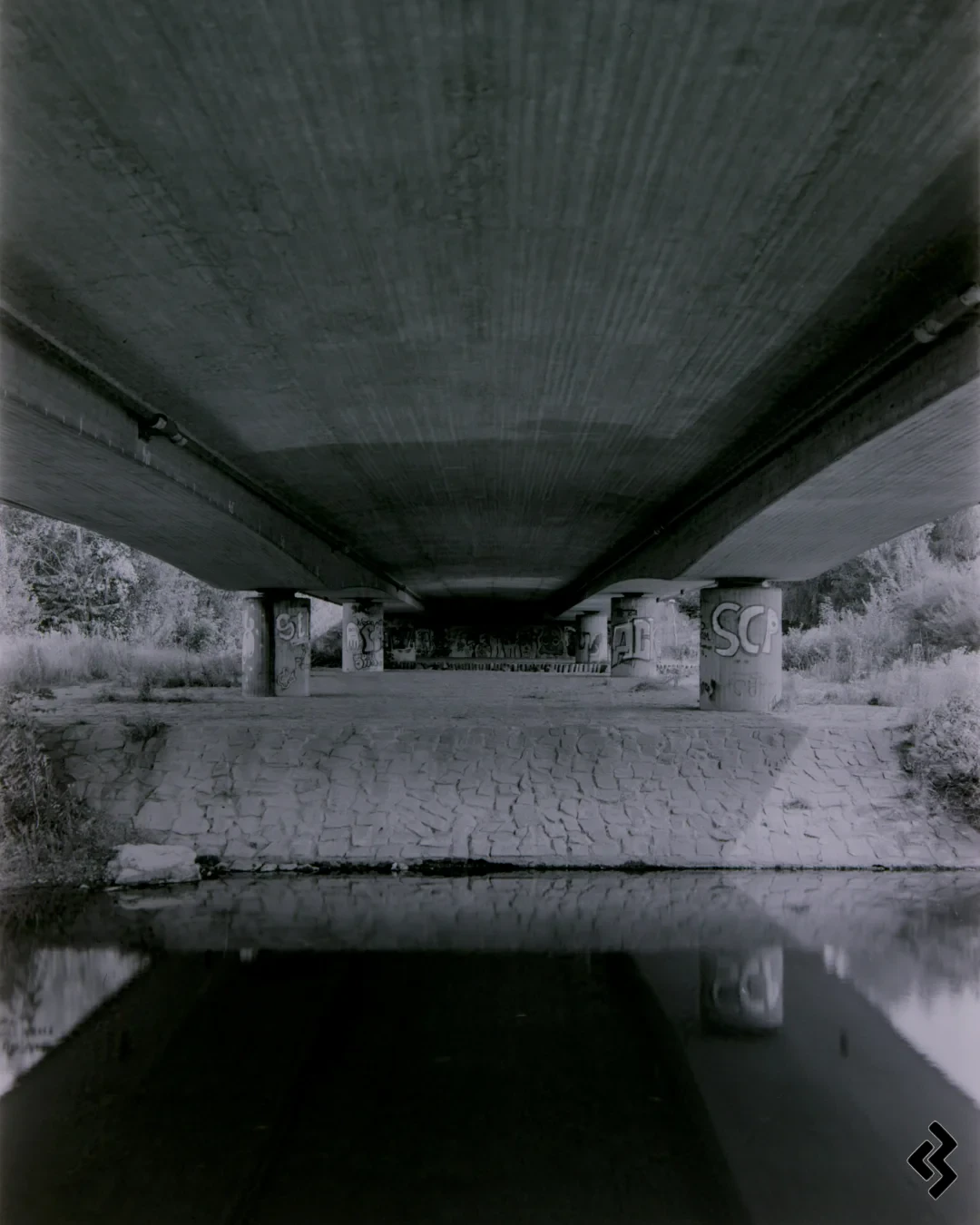Black and white photo taken under a concrete road bridge. The bridge is over a canalized river, the river banks and the ground are paved with stones. The bridge is resting on cylindrical pillars which are covered in graffiti