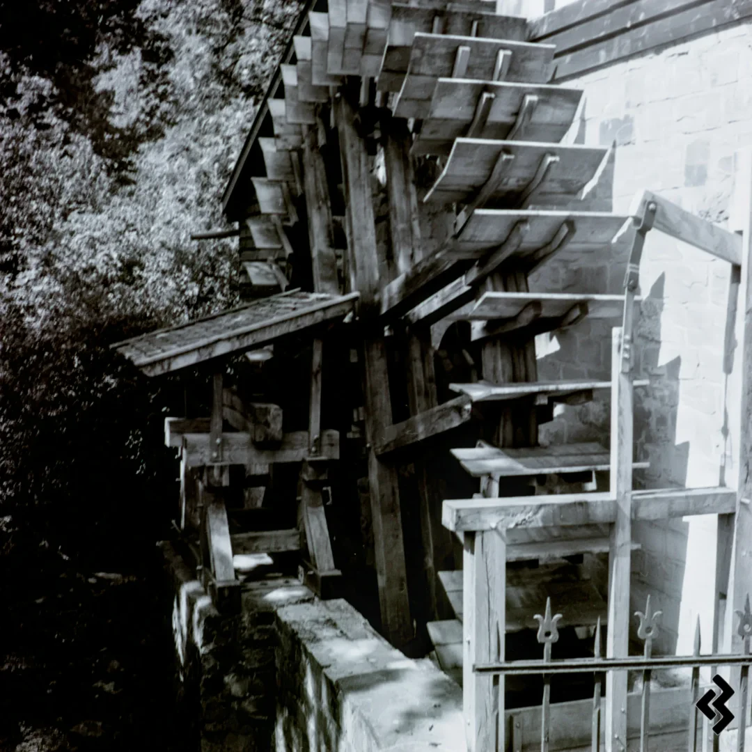 Square format black & white photo of the wooden water wheel of an old watermill. The outer bearing of the wheel has a tine shingled roof and is sitting on a rubblework wall, the other side of the wheel is directly mounted to the buildings wall. In front of the wheel is a gate to control the water flow, it is operated with a lever that reaches into the building. In front of the control gate is a decorative metal fence with tips looking like lillies